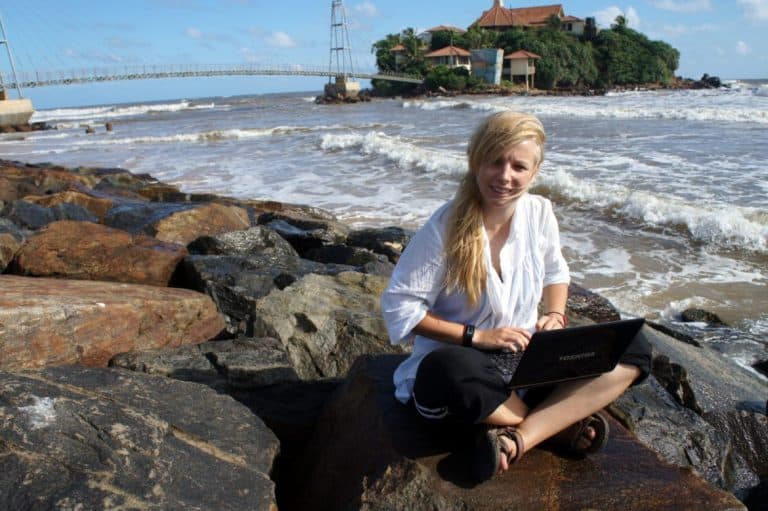 Agness sitting on the stones at the seaside typing on laptop