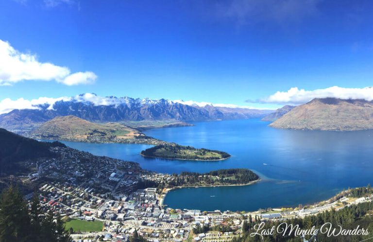 View from mountain top of town by large blue lake with mountains surrounding it.