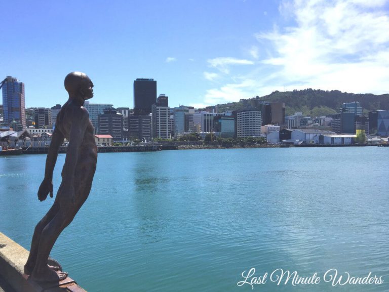Statue of man leaning forward with arms behind him over the water and city buildings behind.