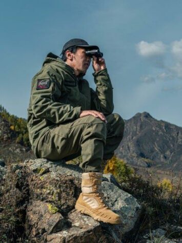 man in boots and jacket looking out binoculars on rock
