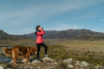 woman in red jacket and dog looking out grass field with binoculars